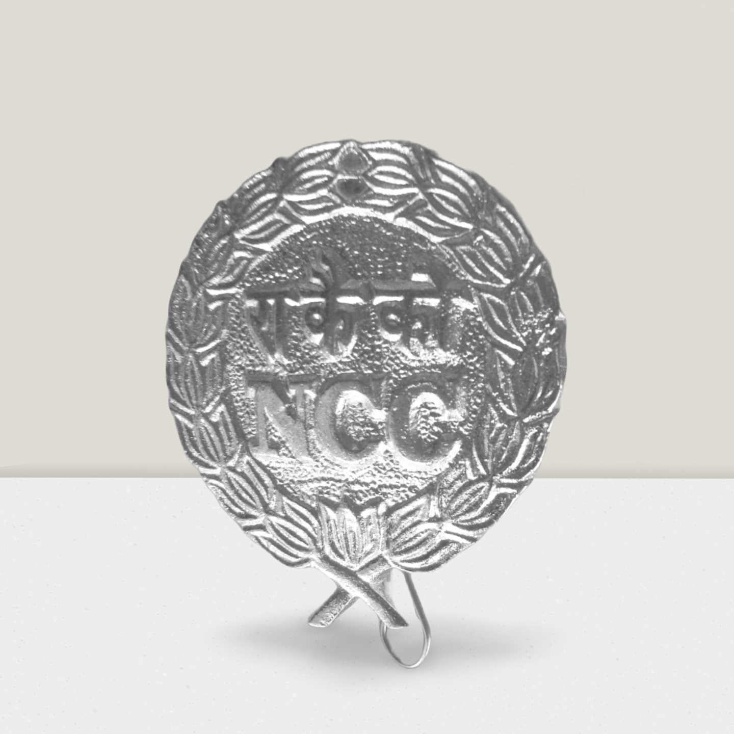 NCC aluminium metal cap badge with embossed ‘NCC’ text and wreath design, lightweight silver-tone finish, featuring crossed torch emblem and attachment loop.